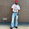 Person wearing a white t-shirt with graphics and text, blue jeans, and a red cap standing against a brick wall.