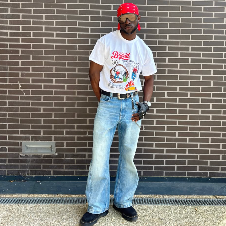 Person wearing a white t-shirt with graphics and text, blue jeans, and a red cap standing against a brick wall.