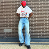 Person wearing a white t-shirt with graphics and text, blue jeans, and a red headscarf standing against a brick wall.