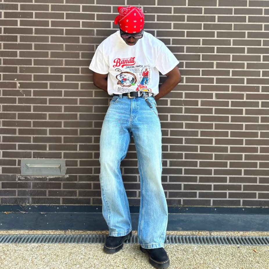 Person wearing a white t-shirt with graphics and text, blue jeans, and a red headscarf standing against a brick wall.