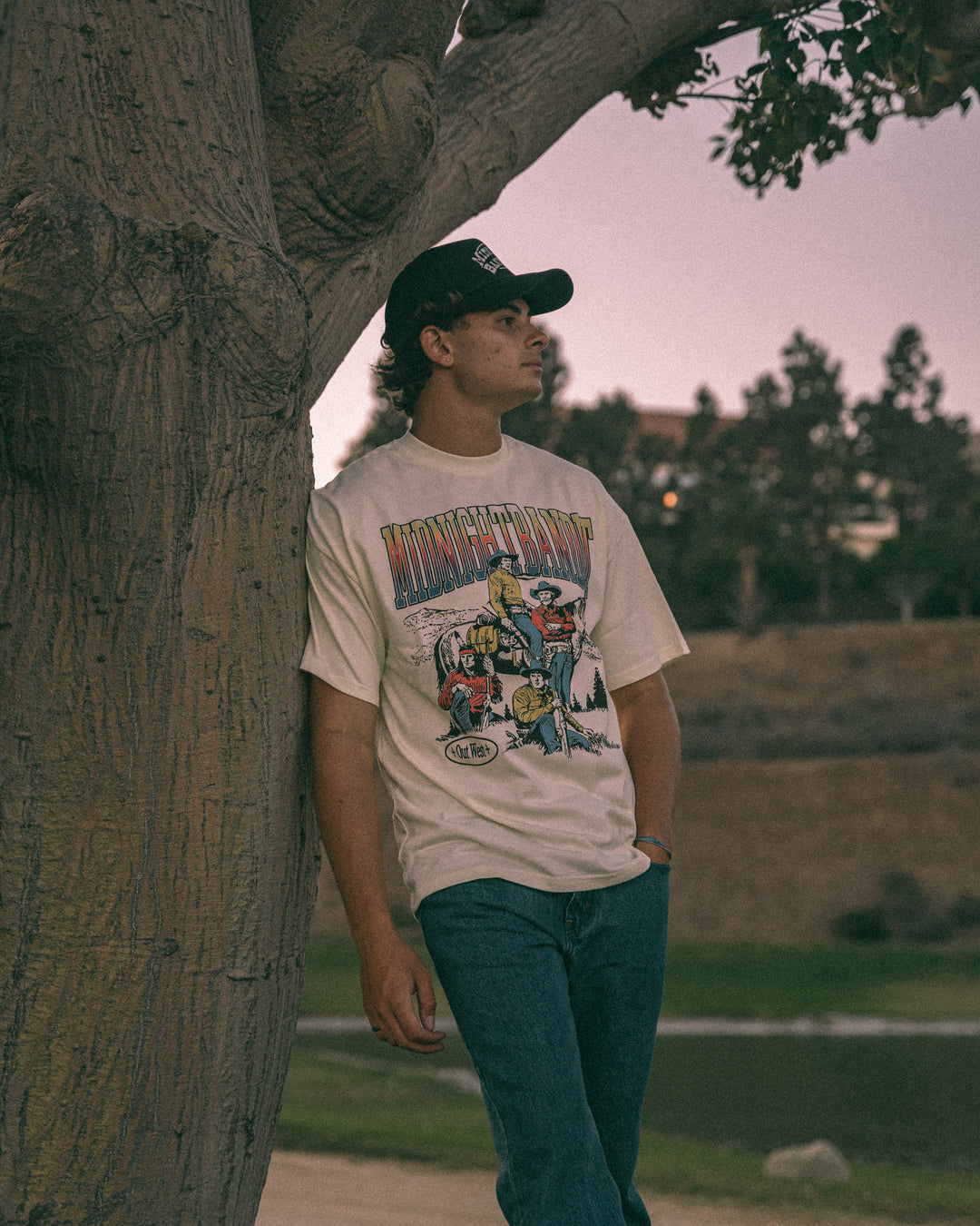 Person wearing a graphic t-shirt leaning against a tree in an outdoor setting