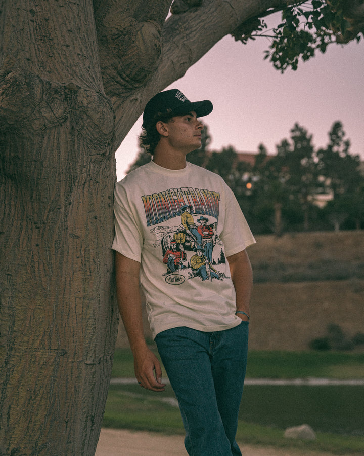 Person wearing a graphic t-shirt leaning against a tree in an outdoor setting