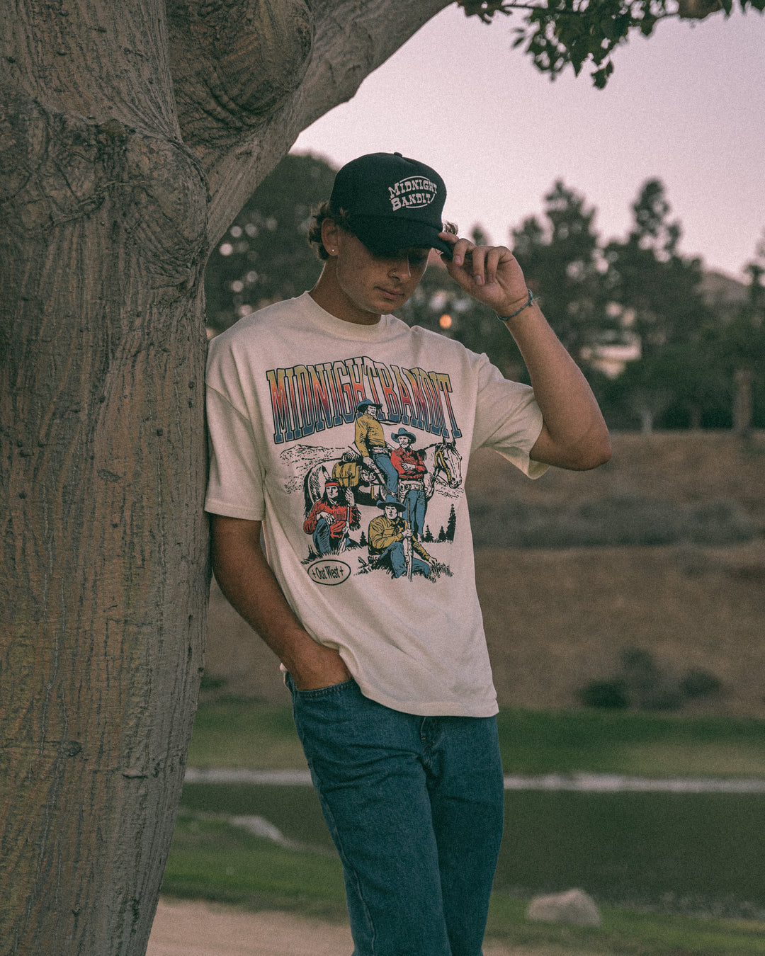 Person wearing a graphic t-shirt and cap leaning against a tree outdoors