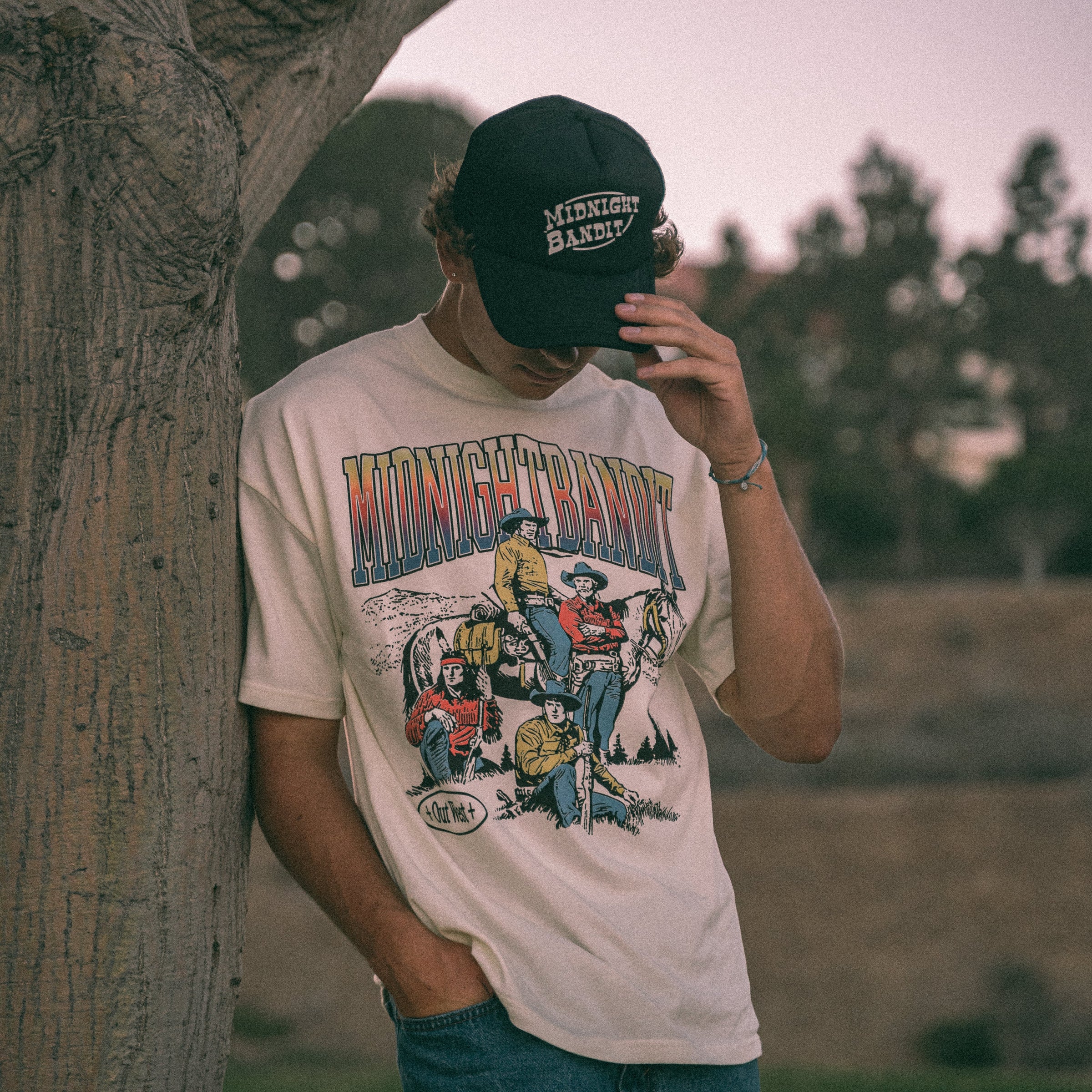 Person wearing a 'Midnight Ranch' t-shirt and cap, standing next to a tree with a blurred natural background