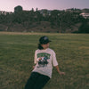 Person sitting on grass wearing a cap and graphic t-shirt with a scenic background